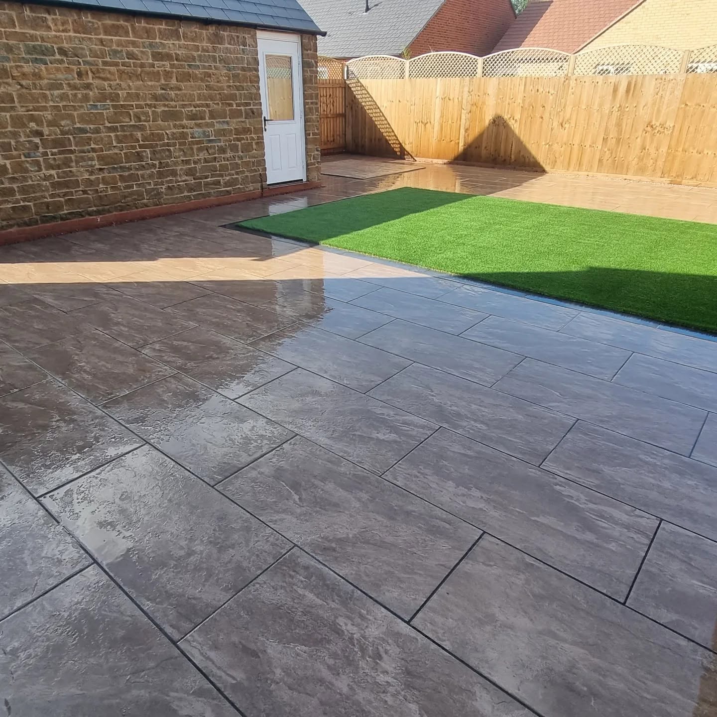 Dark grey paving beside a bright green lawn strip and a new fence