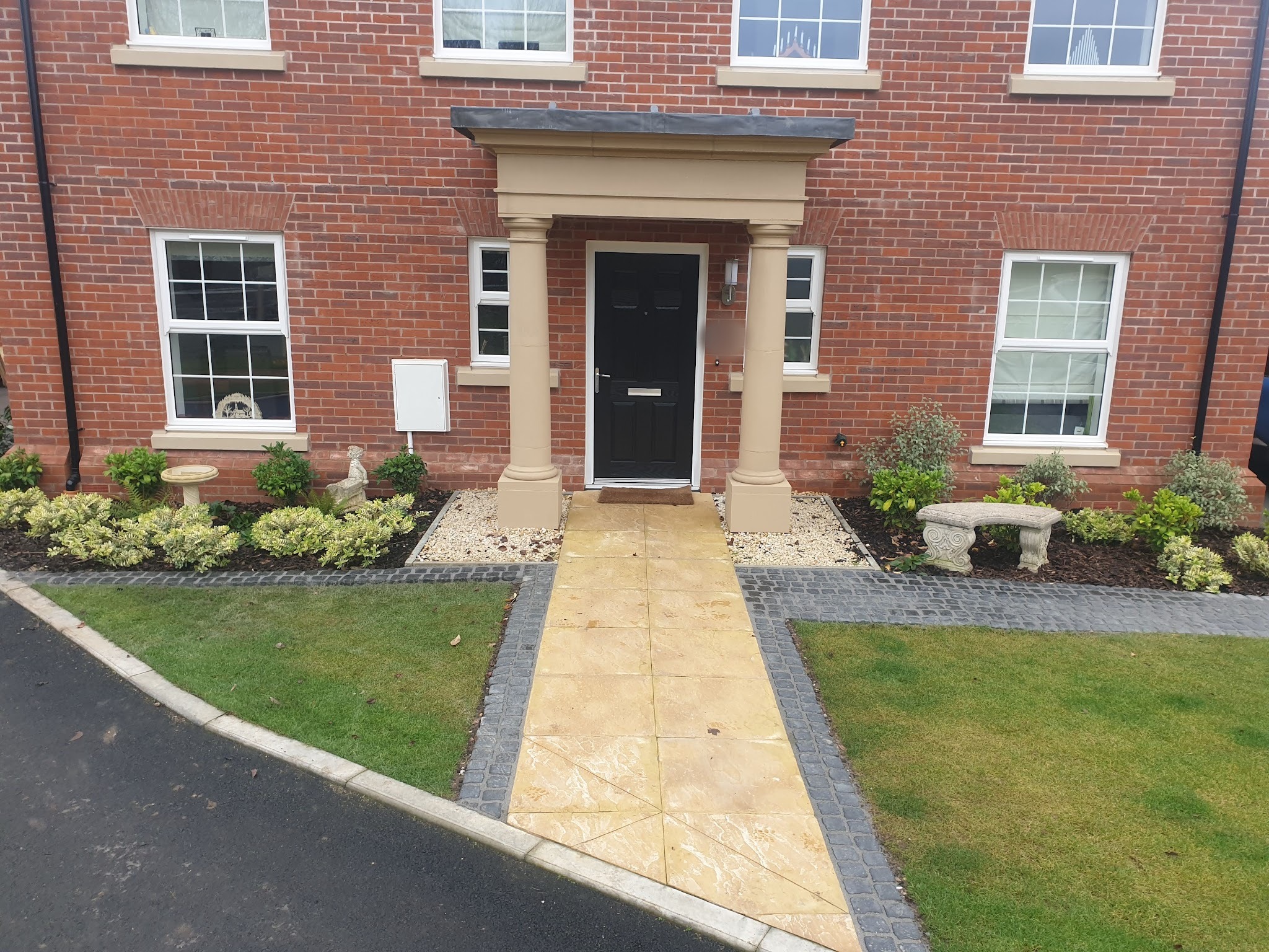 Front garden of a new-build house with a buff paved path, planted borders and lawns
